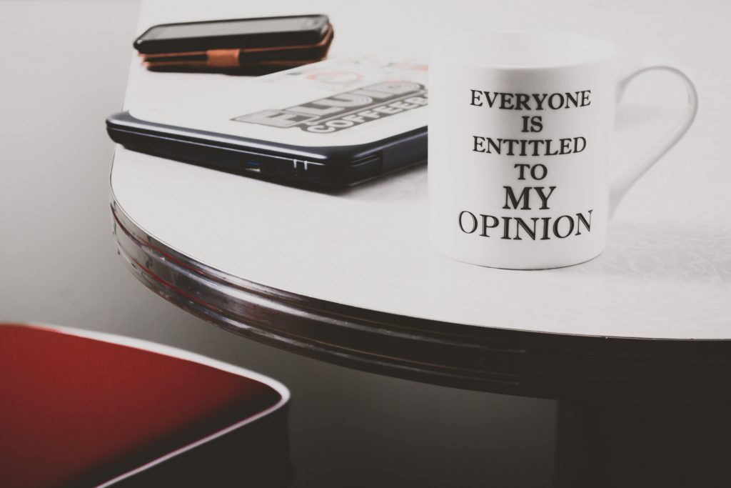 Caneca branca, em cima de uma mesa de madeira branca e preta, com texto em preto que diz "Todos têm direito à minha opinião".